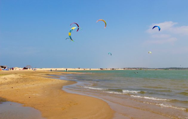 Photo avec en haut un ciel bleu, en bas la plage séparer en deux par la mer et des sportif qui font du kite surf