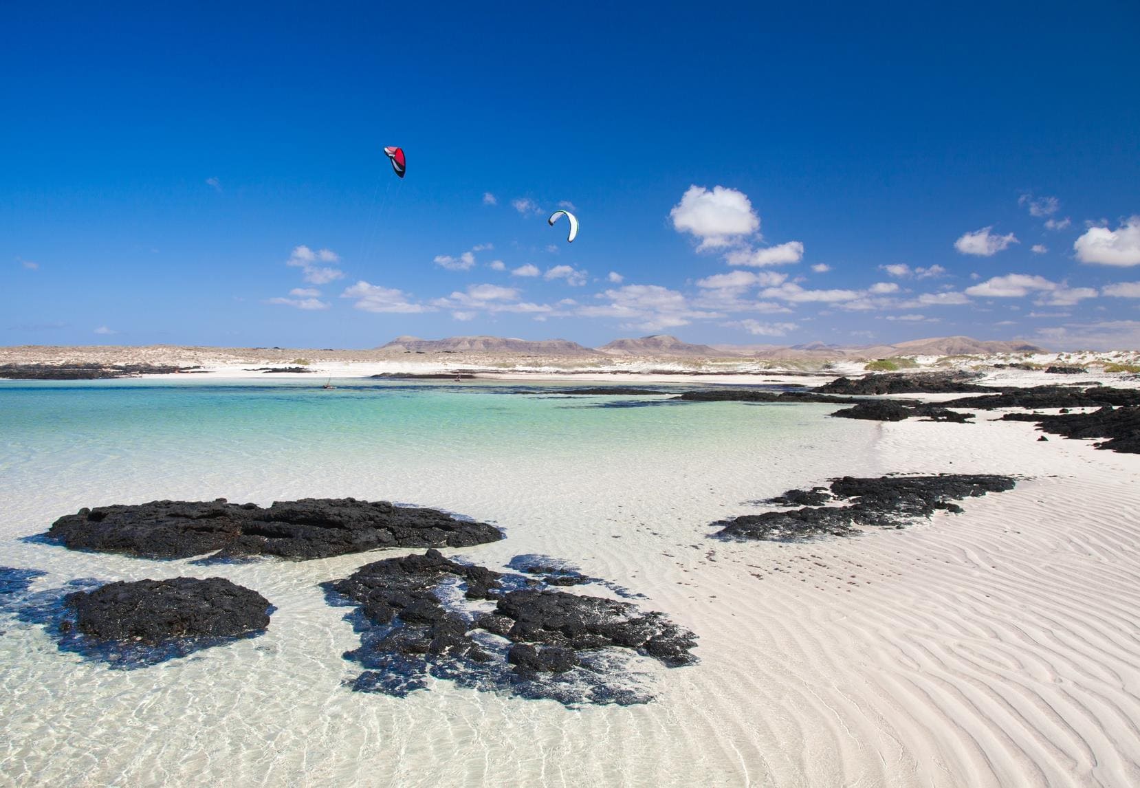 Image avec un ciel bleu en haut, la mer peu profonde en bas avec quelque rocher et des sportif qui font du kite surf.