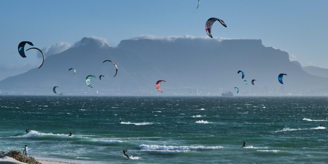 Une photo de sportif qui font du kitesurf, on voit des montagne au loin et la mer en bas de la photo